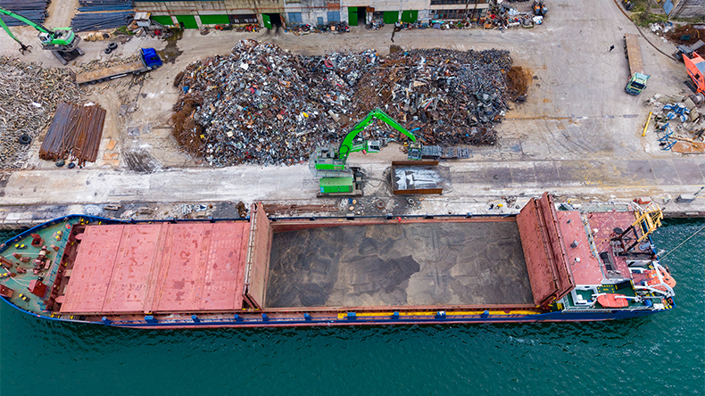 Cargo ship docks at port to unload materials near industrial site with large piles of scrap and storage buildings in the background during day